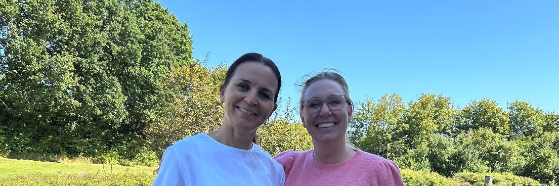 Betina Bruun og Maja Andersen står side om side udendørs på en græsplæne i solskin.
